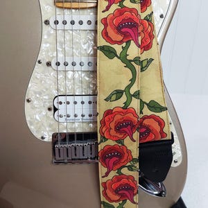 May include: A guitar strap with a repeating floral design of red, orange, and green roses with open mouths and long tongues. The strap is tan with black leather ends and is shown on a gold electric guitar.