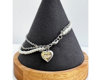 May include: A silver chain bracelet with a heart charm and white pearl accents. The bracelet is displayed on a black velvet stand.