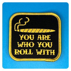 May include: Black embroidered patch with yellow stitching. The patch features a stylized image of a joint with smoke rising from it. The text "You are who you roll with" is embroidered in yellow.