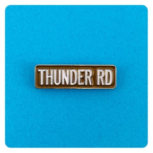 May include: A brown enamel pin with a silver border. The pin reads "THUNDER RD" in white letters.