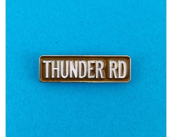 May include: A brown enamel pin with a silver border. The pin reads "THUNDER RD" in white letters.