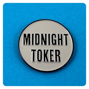 May include: A white enamel pin with a black border and the text "MIDNIGHT TOKER" in black.