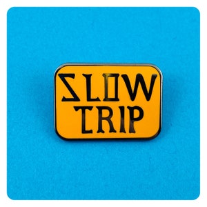 Pode incluir: Um broche de esmalte amarelo com contorno preto e o texto "SLOW TRIP" em preto.