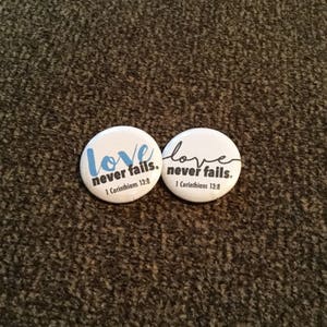 May include: Two white round buttons with the text "love never fails" and "1 Corinthians 13:8". One button has the word "love" in blue script, the other in black script. The buttons are on a textured brown surface.