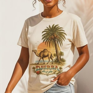Peut inclure: T-shirt crème avec une illustration de scène désertique : chameau, palmiers et le mot "Djerba". Le graphisme utilise des tons chauds. La personne qui porte le t-shirt a des cheveux bouclés et des lunettes de soleil.