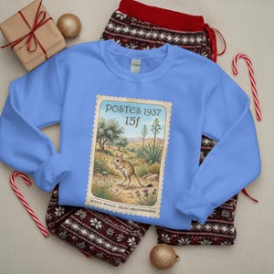 May include: A pale blue sweatshirt with a vintage postage stamp design. The stamp shows a desert landscape with an animal and the text "POSTES 1937 15f". The sweatshirt is placed on red and white patterned pyjama bottoms.