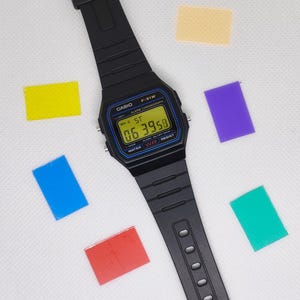 Puede incluir: Reloj digital Casio F-91W negro con una esfera rectangular que muestra la hora. El reloj tiene una correa negra y está rodeado de notas adhesivas rectangulares de colores. La esfera del reloj tiene detalles en azul y amarillo.