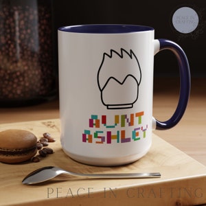 May include: White ceramic mug with a blue rim and handle. The mug features a black outline of a Lego figure head with the text "Aunt Ashley" in colorful Lego bricks.