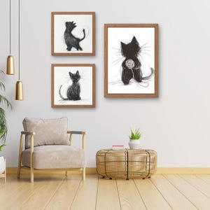 Könnte beinhalten: Drei gerahmte Kunstdrucke mit Katzen. Jeder Druck zeigt eine schwarze Katzenzeichnung auf weißem Hintergrund. Die Rahmen sind in einem warmen Braunton gehalten. Eine Katze sitzt, eine steht und eine spielt mit einem Wollknäuel. Die Kunst wird an einer weißen Wand ausgestellt.