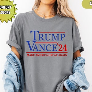 Op de afbeelding: Een grijs t-shirt met de tekst "TRUMP VANCE '24" in blauw en rood. De zin "MAKE AMERICA GREAT AGAIN" is eronder gedrukt. Het shirt is een comfort kleur en is grijs.