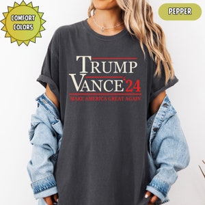 Op de afbeelding: Donkergrijs t-shirt met de tekst "TRUMP VANCE '24" in wit en rood. De zin "MAKE AMERICA GREAT AGAIN" is eronder in het rood gedrukt. Het shirt heeft een relaxte pasvorm en maakt deel uit van de "Comfort Colors" collectie.