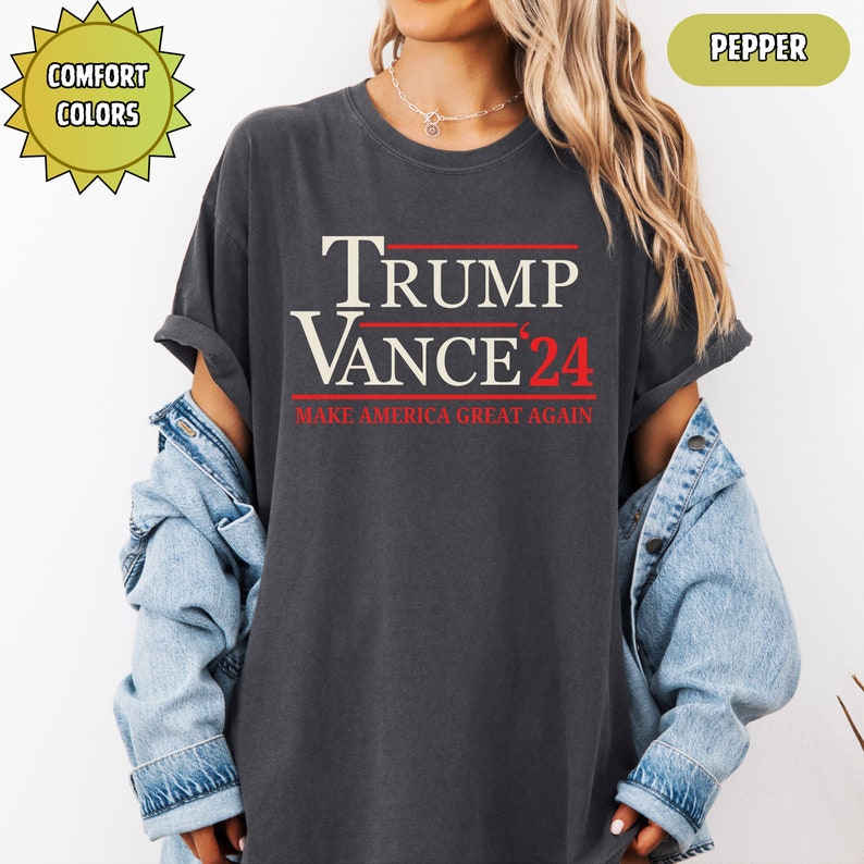 Op de afbeelding: Donkergrijs t-shirt met de tekst "TRUMP VANCE '24" in wit en rood. De zin "MAKE AMERICA GREAT AGAIN" is eronder in het rood gedrukt. Het shirt heeft een relaxte pasvorm en maakt deel uit van de "Comfort Colors" collectie.