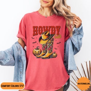 K&ouml;nnte beinhalten: Ein pinkfarbenes T-Shirt mit dem Text "Howdy Fall" in roten und braunen Buchstaben. Das Design zeigt einen Cowboyhut, Stiefel und K&uuml;rbisse.