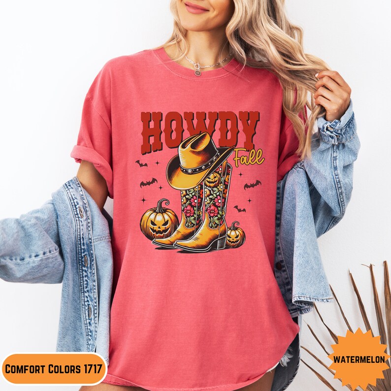 K&ouml;nnte beinhalten: Ein pinkfarbenes T-Shirt mit dem Text "Howdy Fall" in roten und braunen Buchstaben. Das Design zeigt einen Cowboyhut, Stiefel und K&uuml;rbisse.
