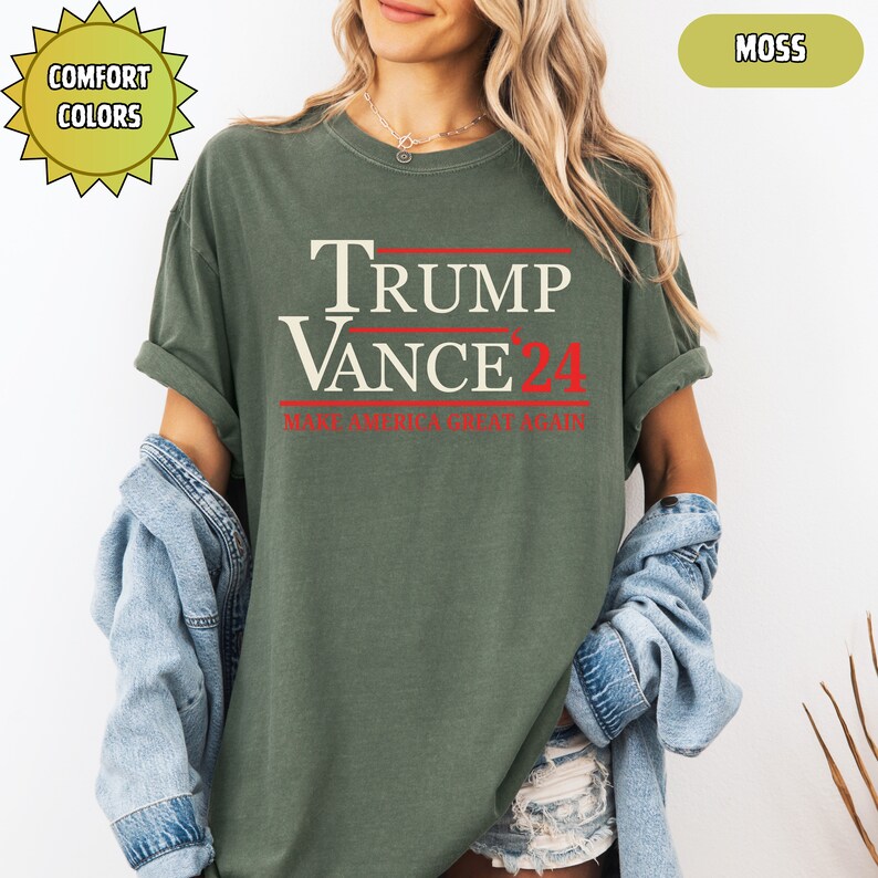 Op de afbeelding: Een mosgroen t-shirt met de tekst "TRUMP VANCE 24" in wit en rood. De zin "MAKE AMERICA GREAT AGAIN" staat er ook in het rood op. Het shirt maakt deel uit van de "Comfort Colors" collectie.