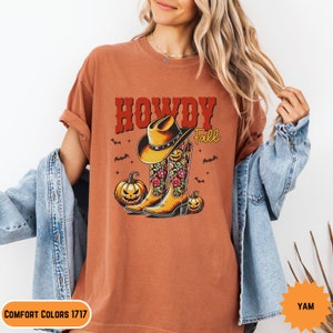 K&ouml;nnte beinhalten: Ein orangefarbenes T-Shirt mit dem Text "Howdy Fall" in Rot und Wei&szlig;. Das Design zeigt einen Cowboyhut auf einem Paar floralen Cowboy-Stiefeln mit K&uuml;rbissen auf beiden Seiten.