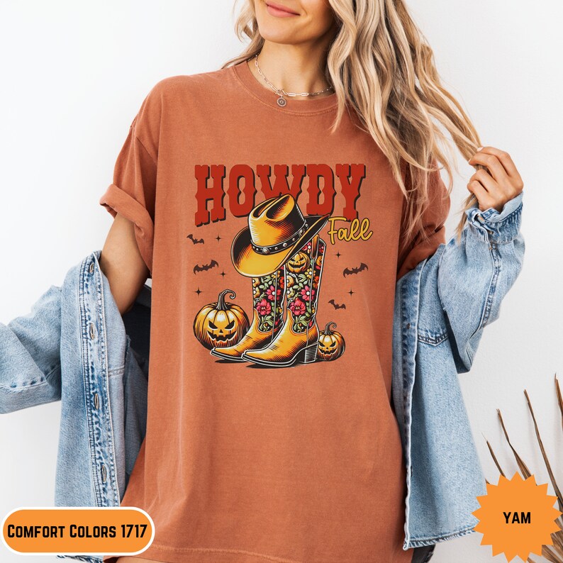 K&ouml;nnte beinhalten: Ein orangefarbenes T-Shirt mit dem Text "Howdy Fall" in Rot und Wei&szlig;. Das Design zeigt einen Cowboyhut auf einem Paar floralen Cowboy-Stiefeln mit K&uuml;rbissen auf beiden Seiten.