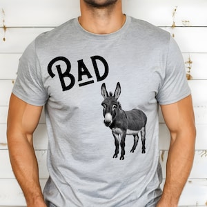 May include: A gray t-shirt with the word "BAD" printed in black, and a black and white donkey illustration below it.