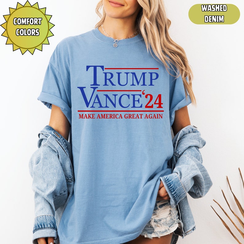 Op de afbeelding: Lichtblauw T-shirt met de tekst "TRUMP VANCE '24" in blauw en rood. De zin "MAKE AMERICA GREAT AGAIN" is eronder gedrukt. Het shirt is gemaakt van een zacht materiaal en is gelabeld met "WASHED DENIM" en "COMFORT COLORS".