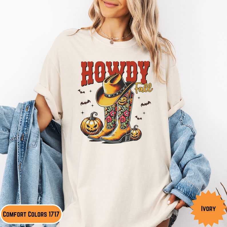 K&ouml;nnte beinhalten: Ein beiges T-Shirt mit dem Text "Howdy Fall" in roten und schwarzen Buchstaben. Das Design zeigt ein Paar Cowboystiefel mit Blumenmustern und einen Cowboyhut sowie K&uuml;rbisse.
