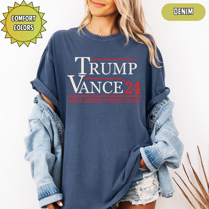 Op de afbeelding: Een denim blauw t-shirt met de tekst "TRUMP VANCE '24" in wit en rood. De zin "MAKE AMERICA GREAT AGAIN" is eronder in het rood gedrukt. Het shirt maakt deel uit van de "Comfort Colors" collectie.
