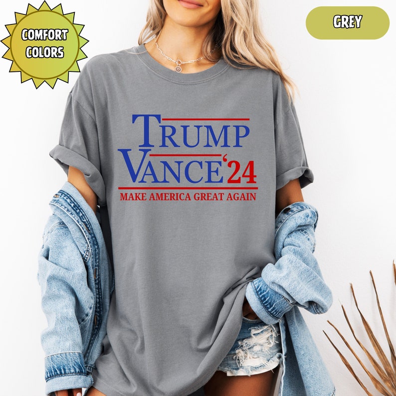 Op de afbeelding: Een grijs t-shirt met de tekst "TRUMP VANCE '24" in blauw en rood. De zin "MAKE AMERICA GREAT AGAIN" is eronder gedrukt. Het shirt is een comfort kleur en is grijs.