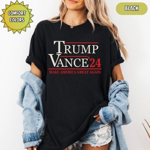 Op de afbeelding: Zwart T-shirt met de tekst "TRUMP VANCE '24" in wit en rood. De zin "MAKE AMERICA GREAT AGAIN" staat onder de namen gedrukt. Het shirt heeft korte mouwen en een ronde hals. De woorden "COMFORT COLORS" staan in een gele ster.