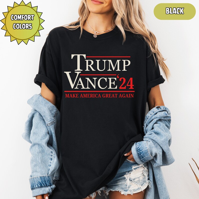 Op de afbeelding: Zwart T-shirt met de tekst "TRUMP VANCE '24" in wit en rood. De zin "MAKE AMERICA GREAT AGAIN" staat onder de namen gedrukt. Het shirt heeft korte mouwen en een ronde hals. De woorden "COMFORT COLORS" staan in een gele ster.