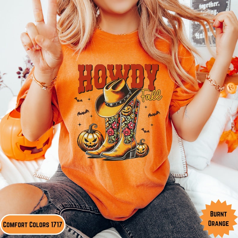 K&ouml;nnte beinhalten: Orangefarbenes T-Shirt mit dem Text "Howdy Fall" und einer Grafik von Cowboystiefeln mit einem K&uuml;rbisdesign.
