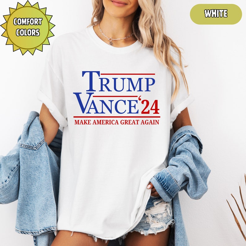 Op de afbeelding: Wit T-shirt met de tekst "TRUMP VANCE '24" in blauw en rood, met "MAKE AMERICA GREAT AGAIN" eronder. Het shirt heeft een ronde hals en korte mouwen. De woorden "Comfort Colors" staan in een gele ster.