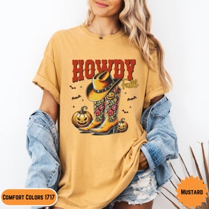 K&ouml;nnte beinhalten: Ein senfgelbes T-Shirt mit dem Text "Howdy Fall" in Rot und einer Grafik von Cowboystiefeln mit Blumendesigns und K&uuml;rbissen.
