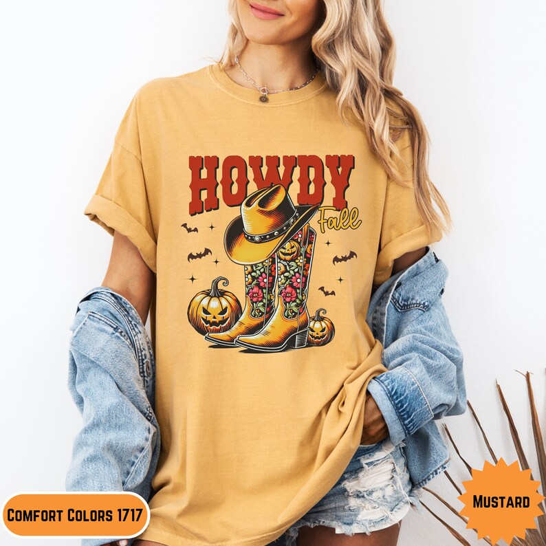 K&ouml;nnte beinhalten: Ein senfgelbes T-Shirt mit dem Text "Howdy Fall" in Rot und einer Grafik von Cowboystiefeln mit Blumendesigns und K&uuml;rbissen.