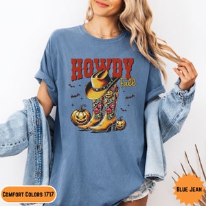 K&ouml;nnte beinhalten: Ein blaues T-Shirt mit dem Text "Howdy Fall" in Rot und Wei&szlig;. Das Design zeigt ein Paar Cowboystiefel mit Blumenmustern und einen Cowboyhut sowie zwei K&uuml;rbisse mit Jack-o'-Lantern-Gesichtern.