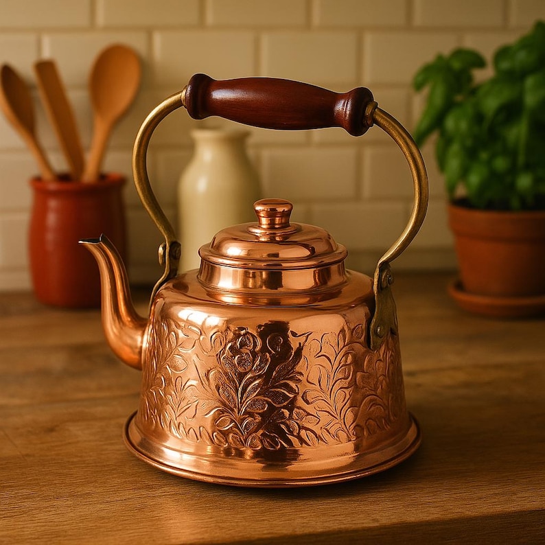 May include: A copper tea kettle with an ornate floral design and a wooden handle. The kettle is sitting on a wooden surface.