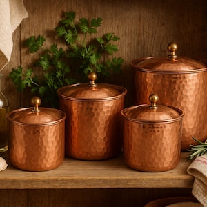 Può includere: Quattro barattoli di rame con texture martellata e pomelli dorati. I barattoli, di diverse dimensioni, sono su una mensola di legno con una bottiglia di vetro, aglio ed erbe. Il colore rame conferisce un aspetto rustico e vintage.