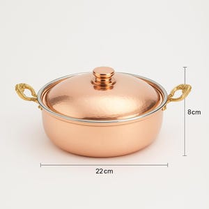 May include: A copper pot with a hammered finish and a lid. The pot has two gold handles and a gold knob on the lid. The pot is 8 inches tall and 22 inches wide.