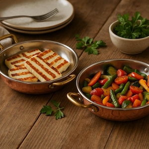 May include: Two copper-colored pans with brass handles, one filled with grilled halloumi cheese and the other with a colorful mix of cooked vegetables. A small bowl of greens and a stack of plates with a fork are also on the wooden table.