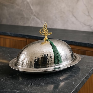 May include: A silver serving dish with a hammered finish and a domed lid. The lid has a gold-colored finial with a green tassel.