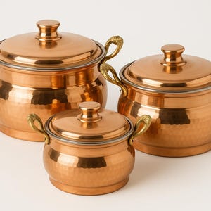 May include: Set of three copper pots with lids and brass handles. The pots are hammered and have a shiny finish.