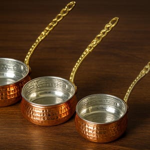 Puede incluir: Tres cafeteras turcas de color cobre con textura martillada e interiores plateados. Cada cafetera tiene un asa decorativa de color dorado. Las cafeteras están dispuestas sobre una superficie de madera oscura.