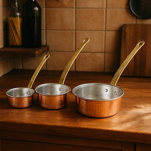 May include: Three copper saucepans with gold handles. The saucepans are nested together, with the smallest saucepan on the left and the largest saucepan on the right.