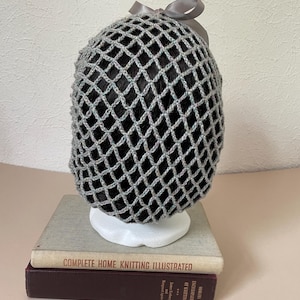 May include: A grey crochet hair net with a diamond-shaped mesh pattern, featuring a small grey satin ribbon bow at the top. The hair net is displayed on a white mannequin head form, which is resting on top of two stacked vintage hardcover books.