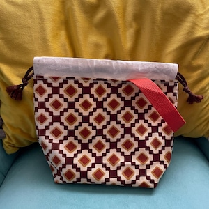 May include: A patterned drawstring bag with a red strap. The bag features a repeating geometric design in burgundy, cream, and gold. The top edge is cream-colored, and the drawstring cords have brown tassels. The bag is sitting on a blue surface.