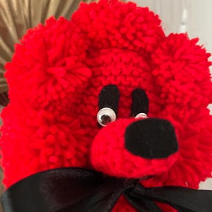 May include: A bright red pom-pom animal figure crafted from yarn, featuring two googly eyes, black felt eyebrows, a black felt nose, and a black satin ribbon tied in a bow around its neck. The texture is fuzzy and rounded with a dense yarn construction.