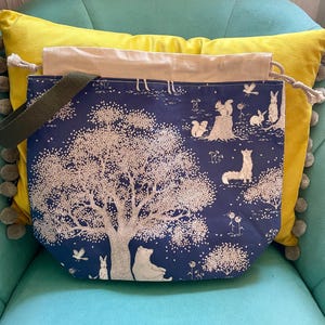 Könnte beinhalten: Eine marineblaue Tragetasche mit einem Waldszenen-Druck. Das Design zeigt einen großen Baum, einen Bären, einen Fuchs, Kaninchen und Eichhörnchen. Die Tasche hat einen grünen Riemen und einen beigen Kordelzugverschluss. Die Tasche liegt auf einer türkisfarbenen Oberfläche.
