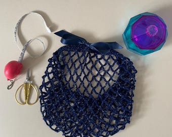 May include: A dark blue crocheted net bag with a matching ribbon bow at the top, accompanied by a red retractable measuring tape, gold-toned metal embroidery scissors, and a faceted teal and purple glass container with a matching lid.