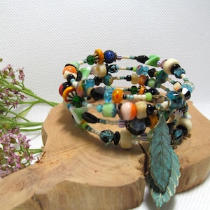 May include: A multi-strand wrap bracelet with colorful beads in shades of blue, green, orange, and cream. A teal leaf charm hangs from the bracelet. The bracelet rests on a piece of wood, with purple flowers in the background.