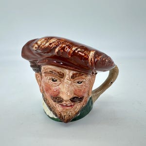 May include: A ceramic mug shaped like a man's face, featuring a brown beret, a mustache, and a beard. The mug has a handle and is painted with realistic details, including skin tones and facial features. The mug is a unique decorative item.