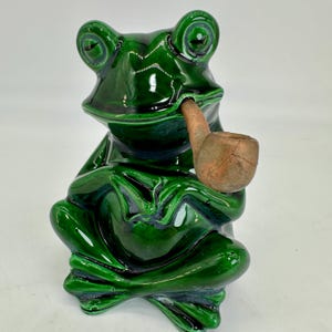 Könnte beinhalten: Glänzende, smaragdgrüne Keramikfroschfigur, sitzend mit gekreuzten Beinen. Der Frosch hält eine braune Pfeife im Mund. Die Figur hat große, ausdrucksstarke Augen und eine detaillierte, strukturierte Oberfläche.