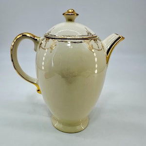 May include: Cream-colored ceramic teapot with a lid and gold trim. The handle, spout, and lid finial are accented with gold. Delicate gold floral patterns adorn the lid and body, creating an elegant vintage aesthetic.